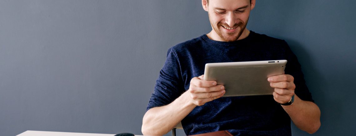 Portrait of bearded man smiling and using pad at desktop against of grey wall